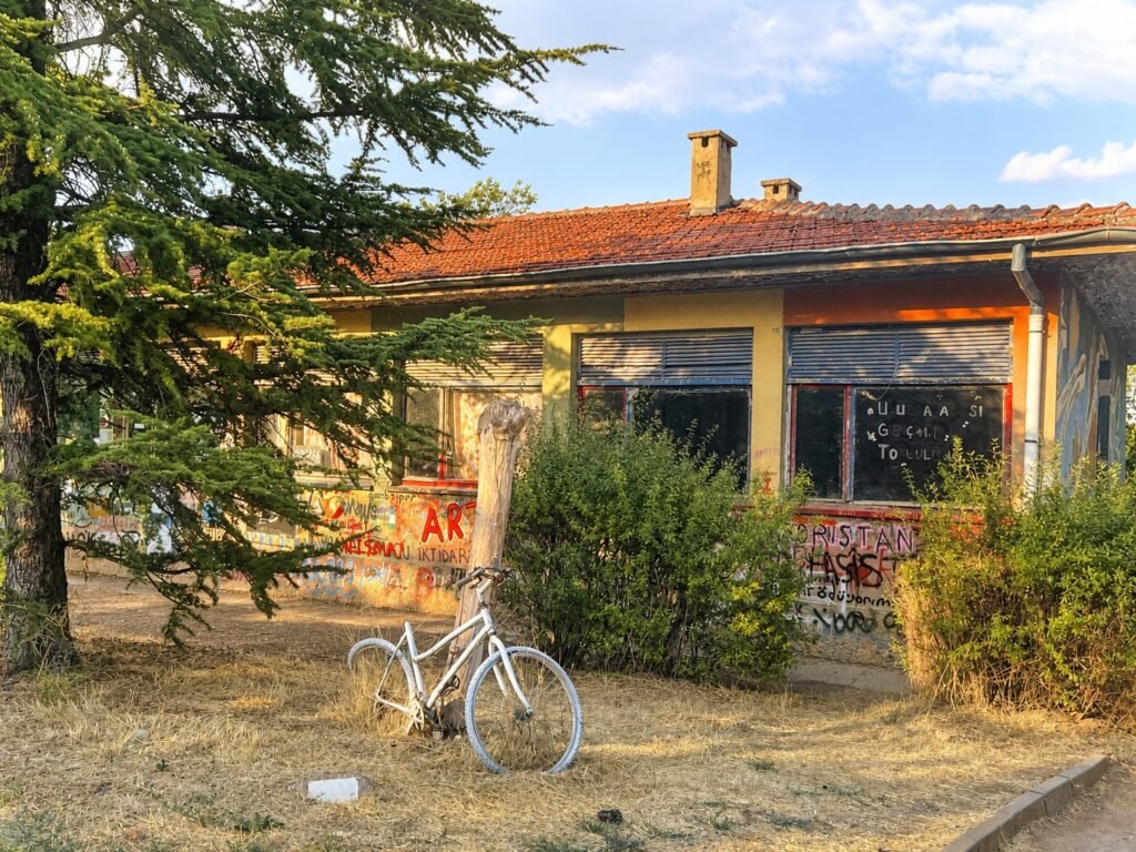 White bicycle leaning against a wooden post outside a graffiti-covered building on METU campus, dry grass in foreground.
