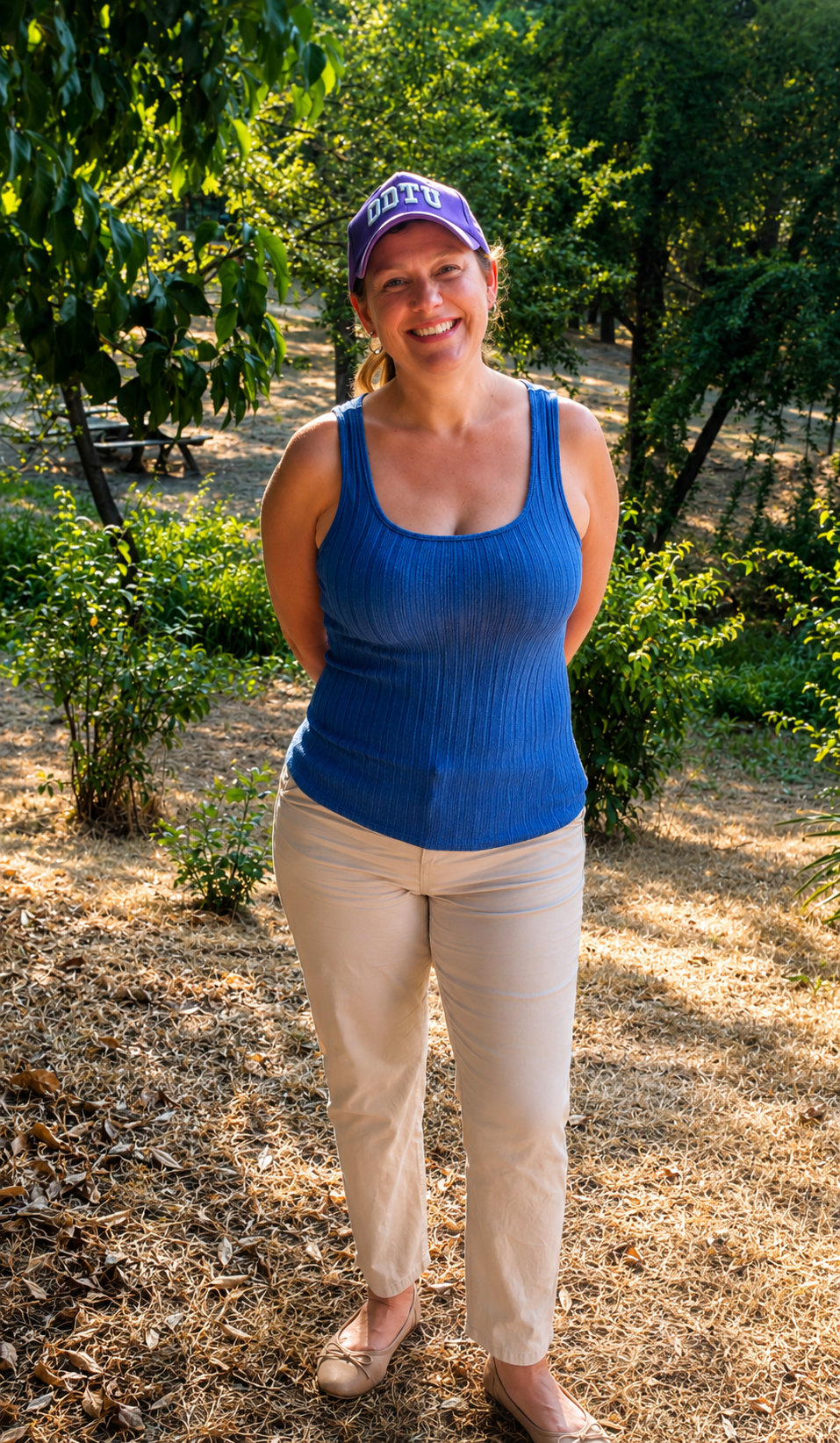 Tiffles smiling outdoors in Ankara wearing a purple ODTÜ cap, blue sleeveless top, beige pants, and ballet flats surrounded by trees.
