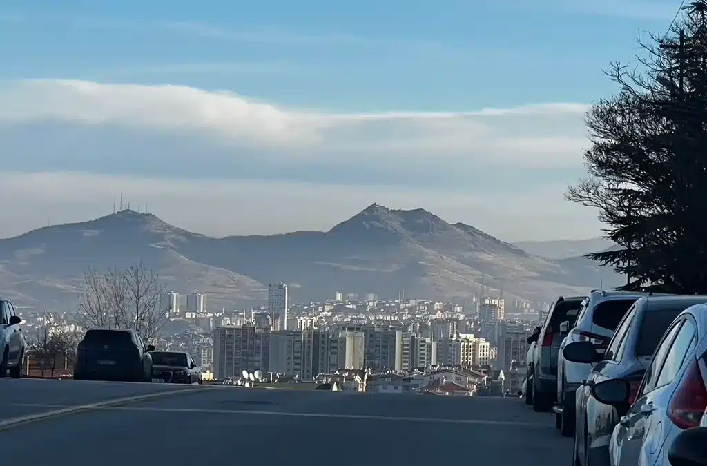A residential street lined with parked cars opens onto Ankara’s apartment buildings and rugged hills beneath a blue sky.