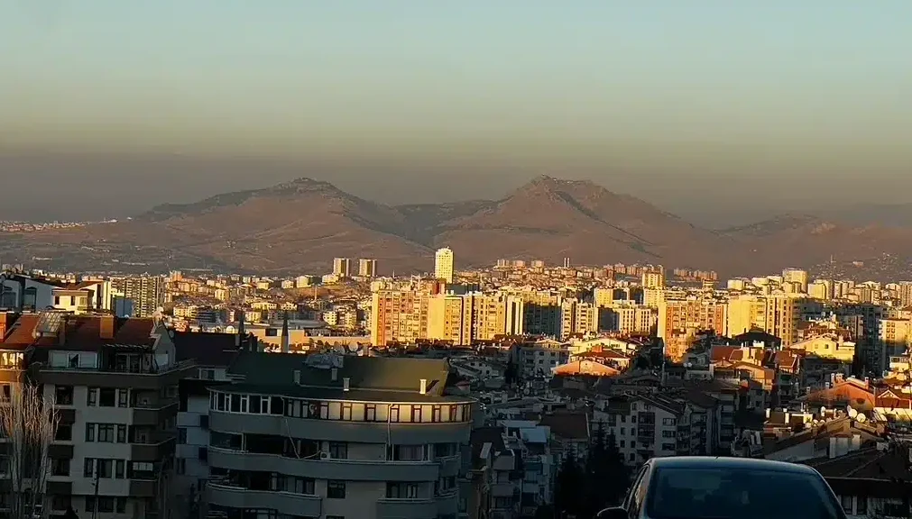 Golden evening sunlight illuminates Ankara's apartment buildings beneath distant hills and a clear sky.
