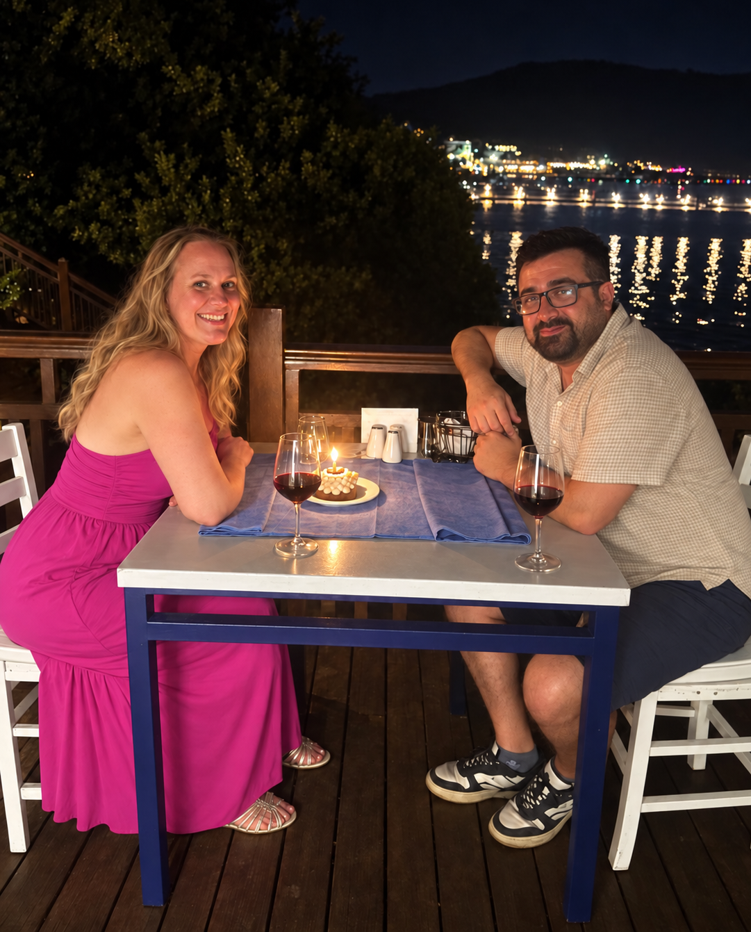 Tiffles and Bora seated beside the water at night with wine and a candlelit dessert, with coastal lights reflected in Torba Bay.
