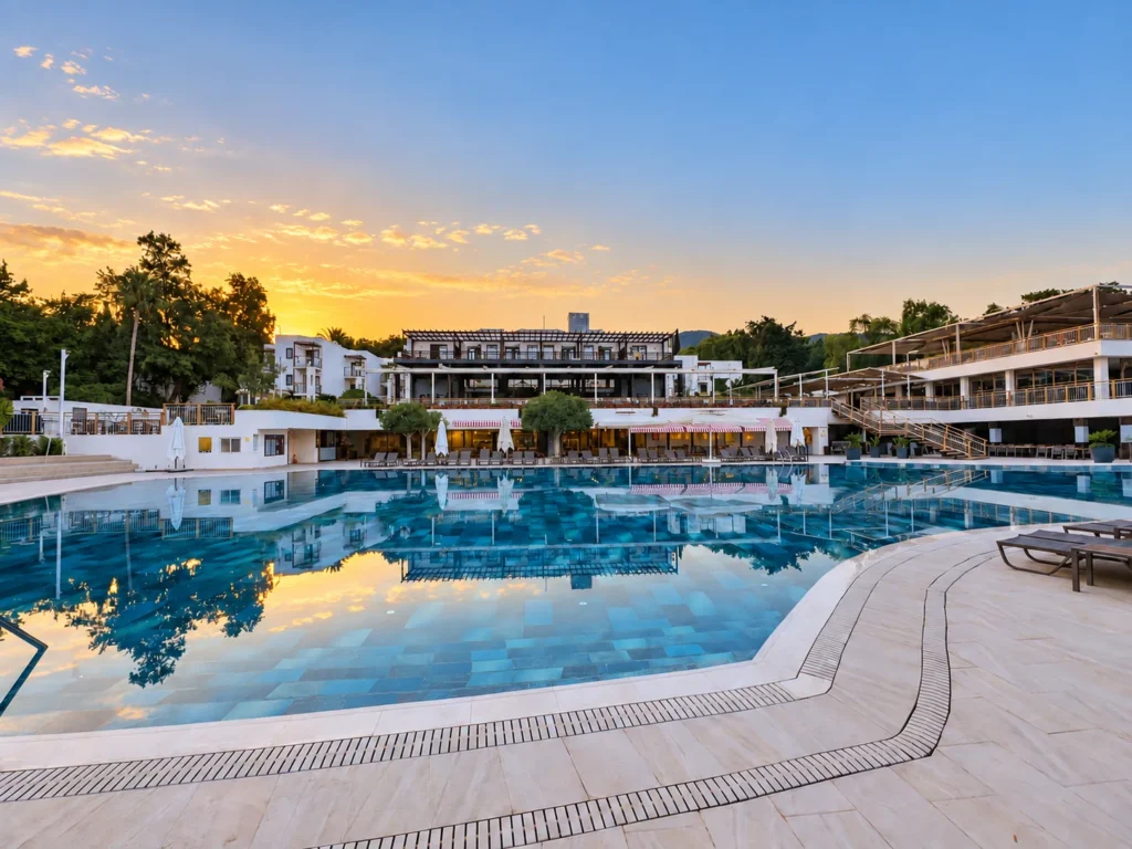 Large resort swimming pool reflecting white buildings, trees, and a softly colored morning sky