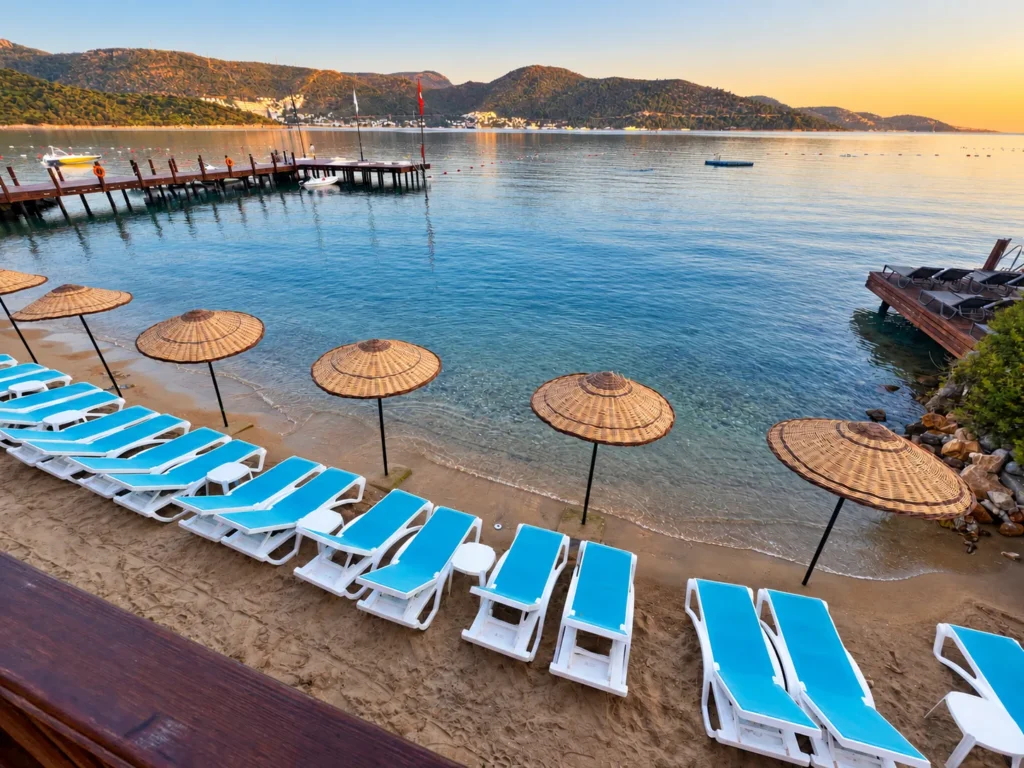 Row of blue and white beach loungers with straw parasols beside a calm Torba Bay in Bodrum.