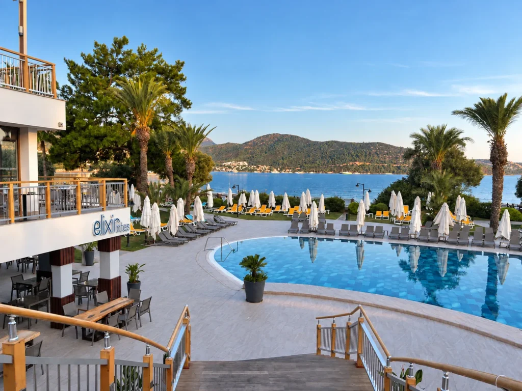 Elevated view over a curved resort pool, palm trees, white umbrellas, and Torba Bay in Bodrum.