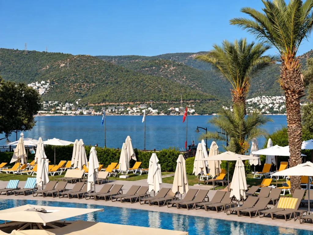 White beach umbrellas and loungers beside a blue Bodrum bay with hillside homes in the distance