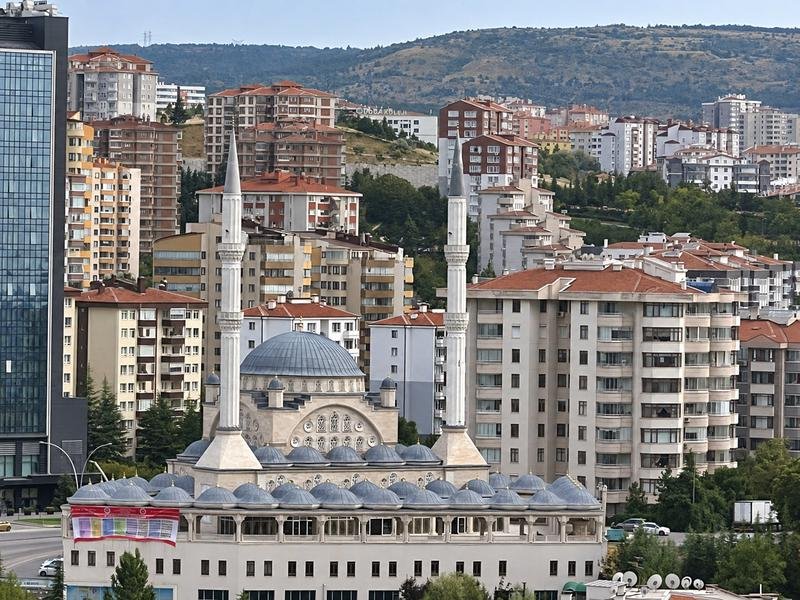 Mosque with twin minarets and a gray dome among Ankara apartment buildings, with hills in the background.