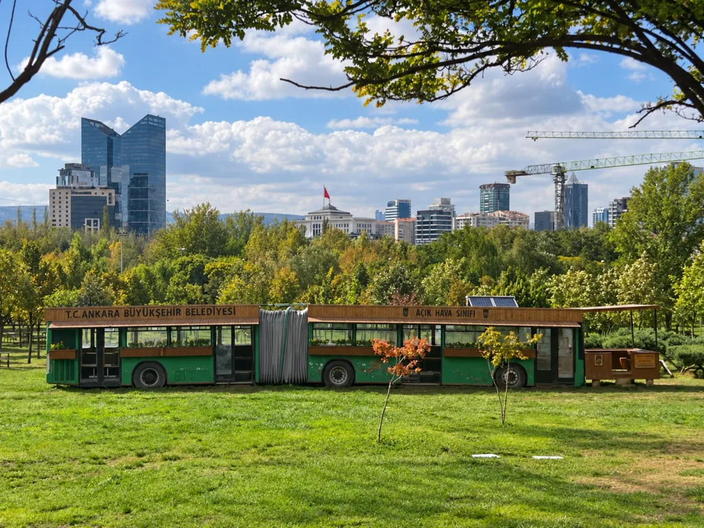 A green articulated bus marked as an open-air classroom stands on the lawn below Ankara’s skyline.