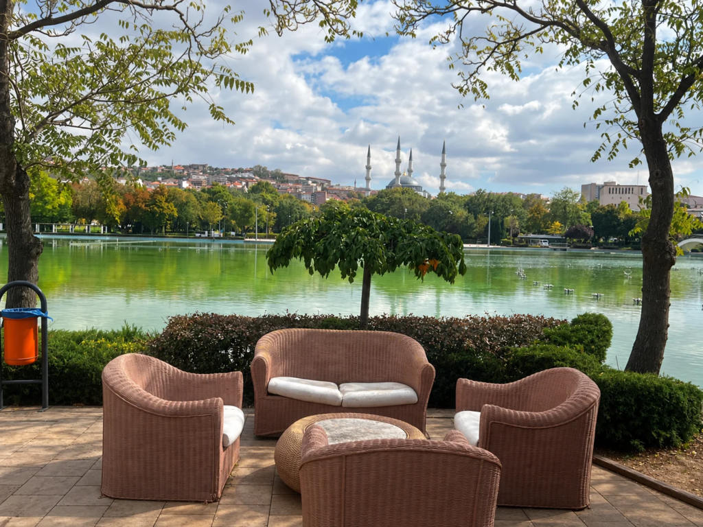 Wicker chairs and a loveseat beside the pool in Gençlik Parkı, with trees, a mosque, and Ankara’s skyline beyond.