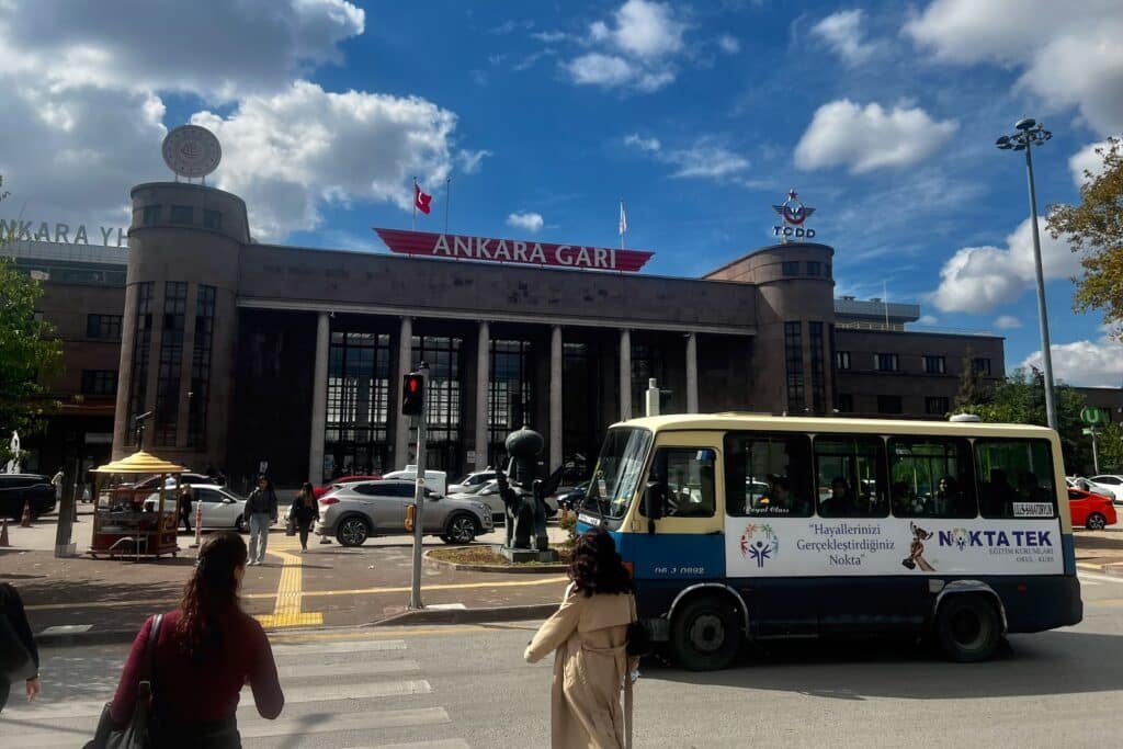 Historic Ankara Gar railway station beneath a blue sky with clouds, with pedestrians and vehicles in front.