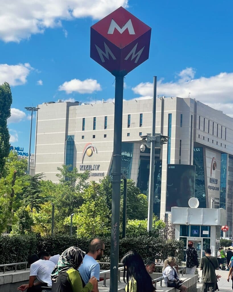 Red Ankara Metro sign above the station entrance in Kızılay, with pedestrians and surrounding buildings.