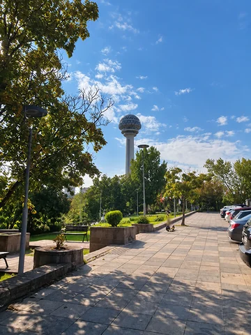 Atakule rises above trees beside a paved walkway and parked cars at Botanik Parkı.