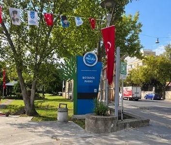 A blue Botanik Parkı entrance sign stands beside trees and Turkish flags near the road.