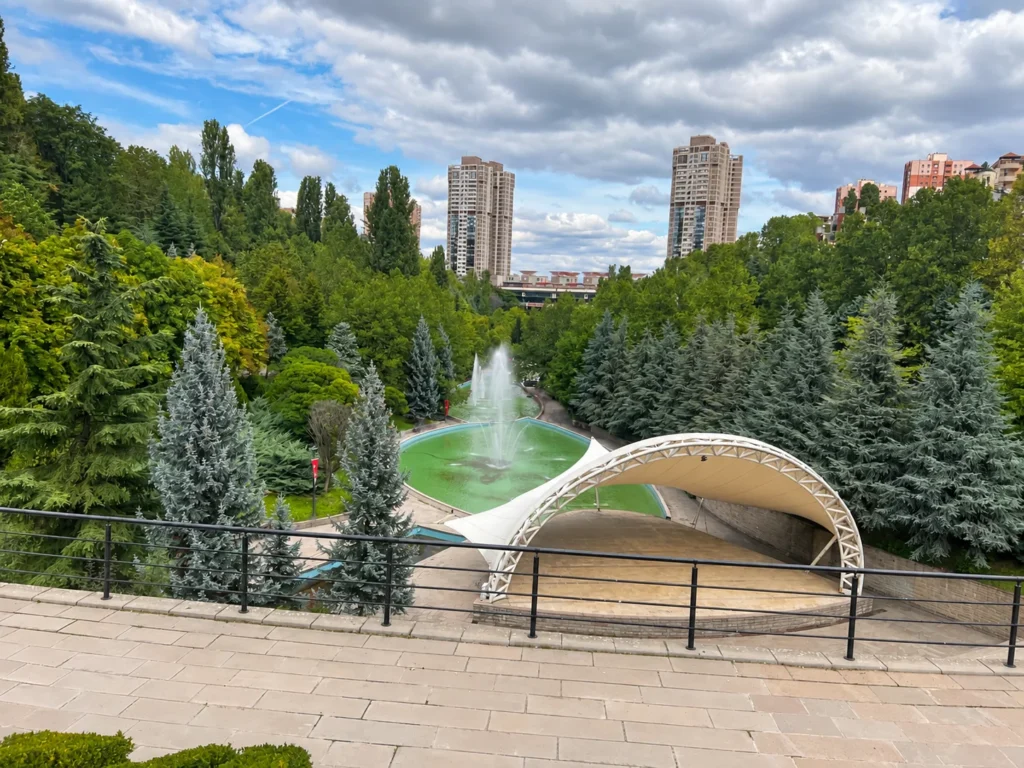 Fountain pools and an amphitheater canopy below a railing, surrounded by trees and towers in Dikmen Vadisi.