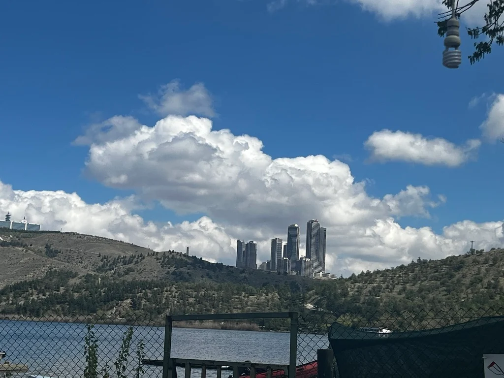 Tall apartment towers rise beyond wooded hills and Lake Eymir, seen through a lakeside fence beneath large white clouds.