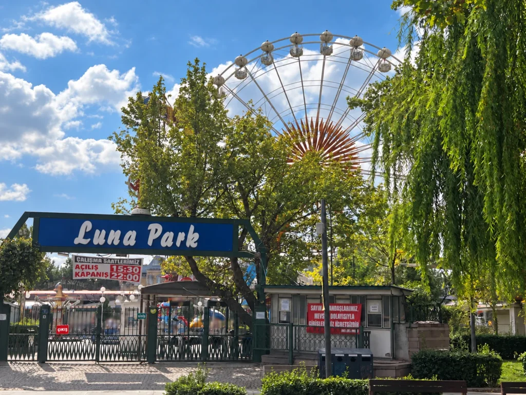 A Luna Park entrance sign stands beneath a Ferris wheel partly framed by trees and a blue, cloud-filled sky.