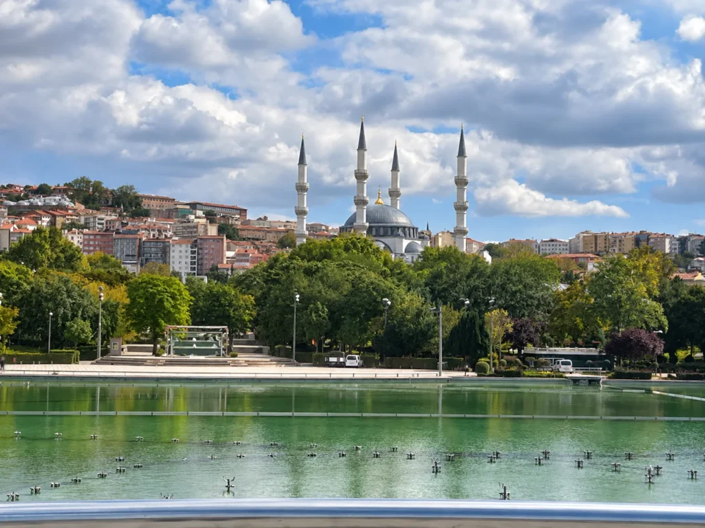 A domed mosque with four minarets rises above trees and city buildings beyond Gençlik Parkı's green pool.