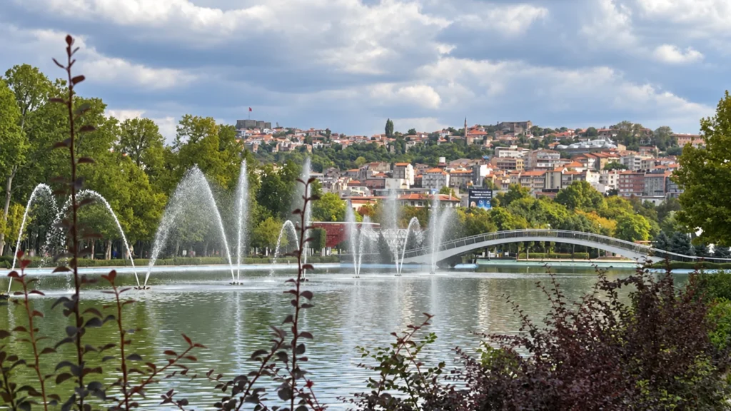 Fountains spray across Gençlik Parkı's pool beside an arched pedestrian bridge, with hillside buildings beyond.