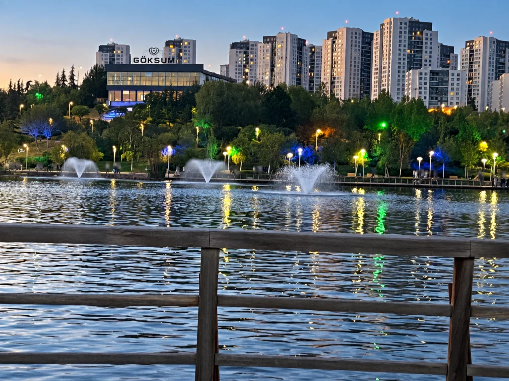 Fountains spray across Göksu Lake beneath apartment towers, seen behind a wooden lakeside railing.
