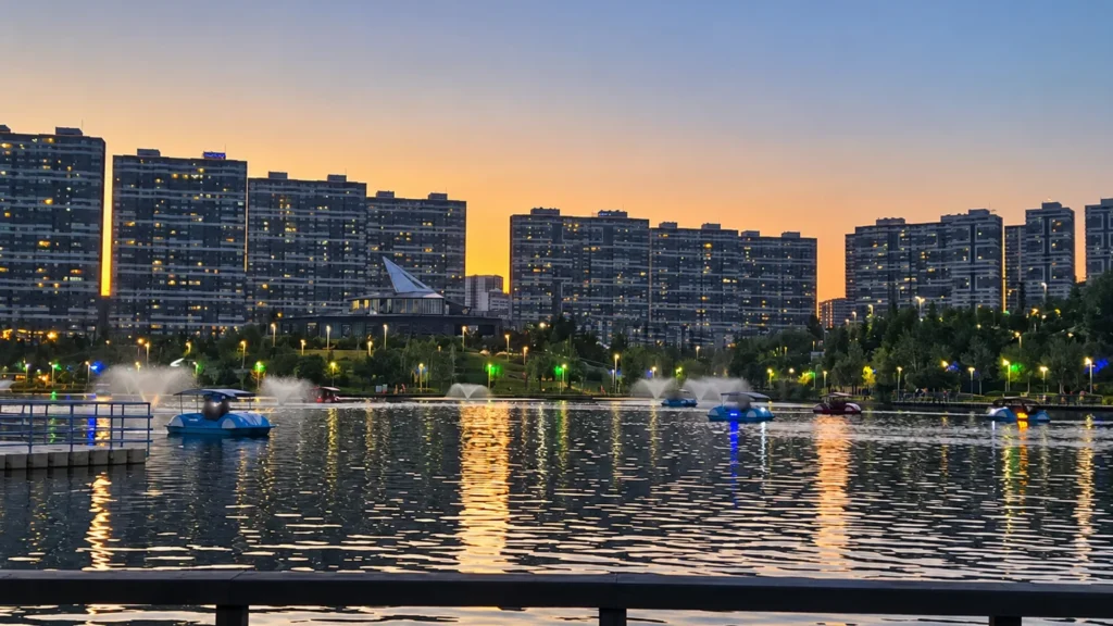 Boats and fountains dot Göksu Lake beneath a dusk sky, with apartment blocks and reflections across the water.