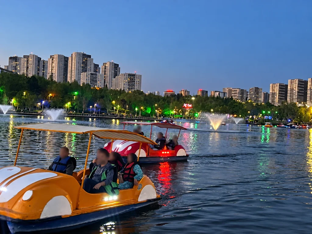 Orange and red leisure boats carry visitors across Göksu Lake at dusk, with fountains and apartment buildings beyond.