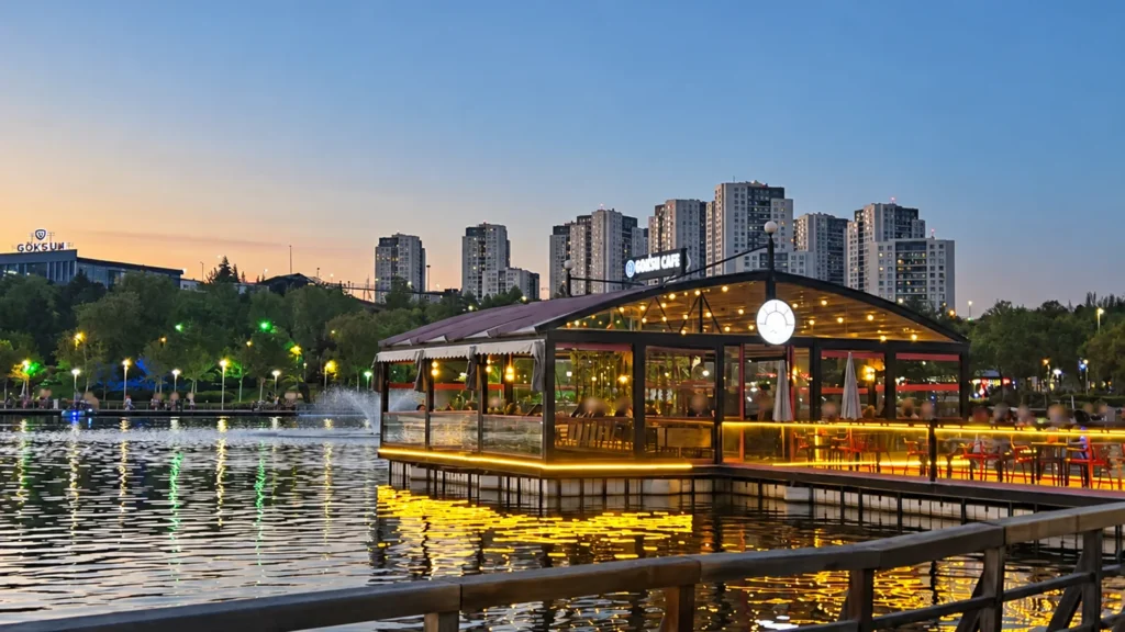 An illuminated café stands beside Göksu Lake, casting golden reflections on the water beneath a dusk skyline.