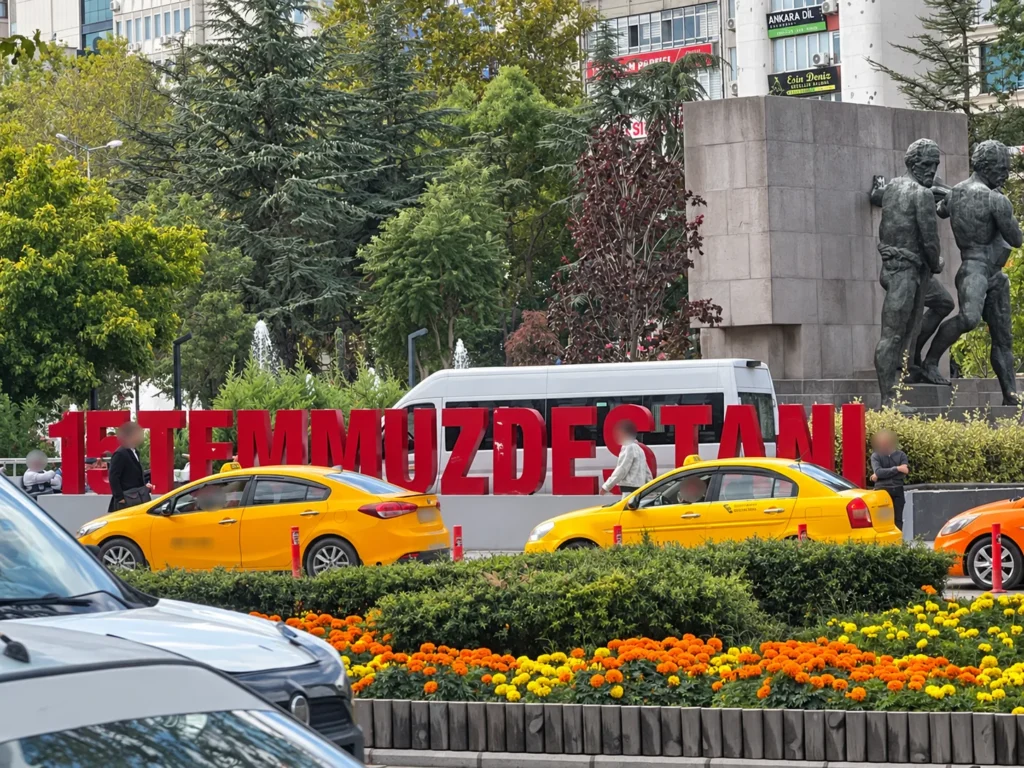 Yellow taxis pass red lettering and the sculptural monument beside Güvenpark, with marigolds in front.