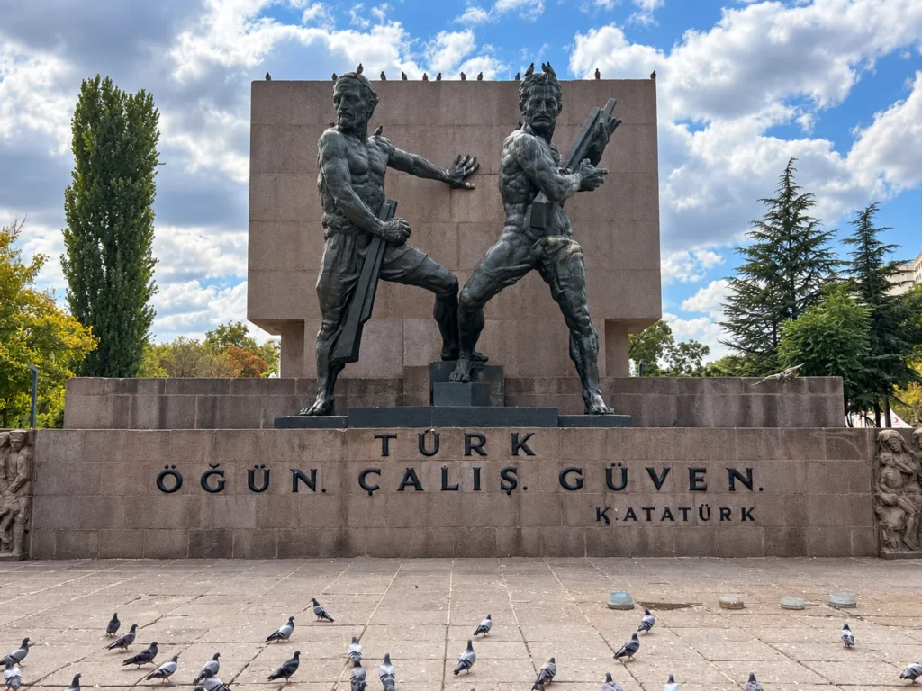 Two bronze figures stand against a stone monument above a Turkish inscription, with pigeons on the paving below.