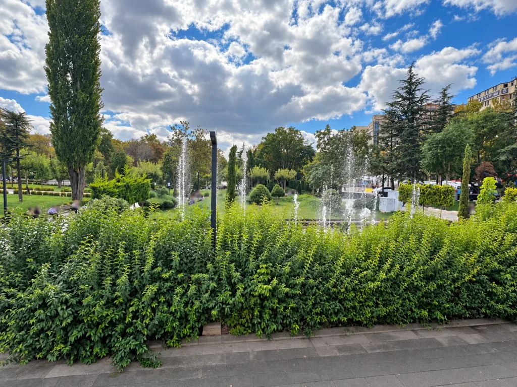 Fountain jets rise behind a leafy hedge, with lawns and tall trees in Güvenpark.