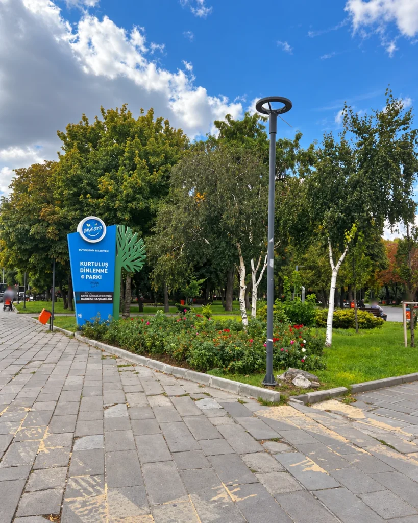 Blue Kurtuluş Parkı entrance sign beside roses, trees, and a paved walkway in Ankara.