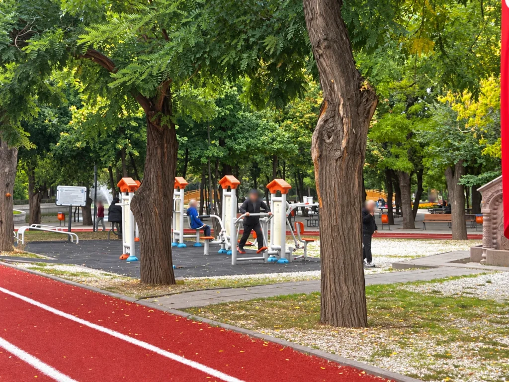 White and orange outdoor exercise machines stand beneath trees beside a red running lane in Kurtuluş Parkı.