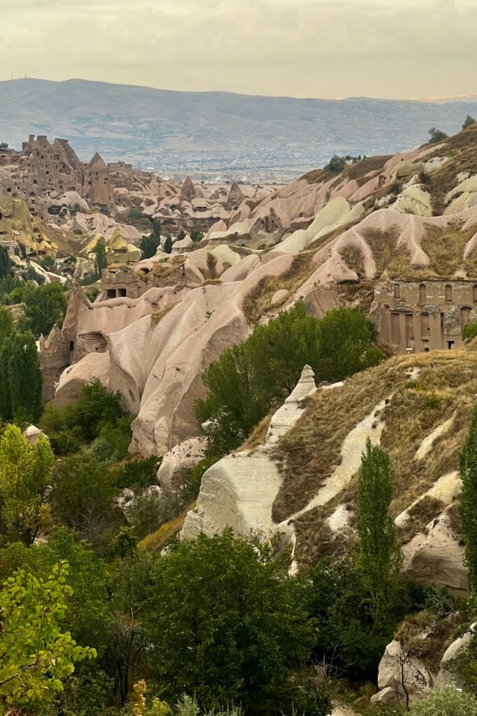 Wide view of Pigeon Valley with carved rock formations, green trees, and distant mountains in Cappadocia, Türkiye