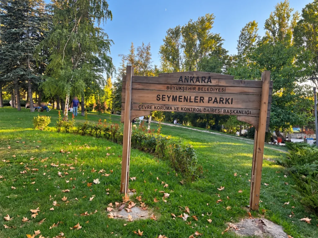 A wooden Seymenler Parkı sign stands on a grassy slope surrounded by trees and fallen leaves.