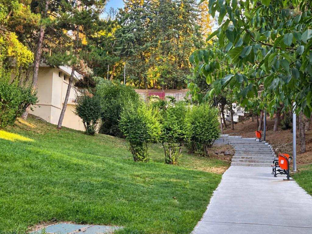 A paved path leads past benches and clipped shrubs toward steps on a grassy hillside in Seğmenler Parkı.