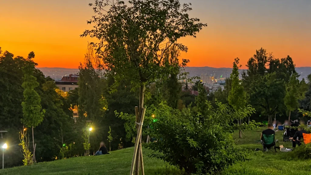 An orange sunset glows above Ankara’s distant skyline beyond trees and a sloping lawn in Seğmenler Parkı.